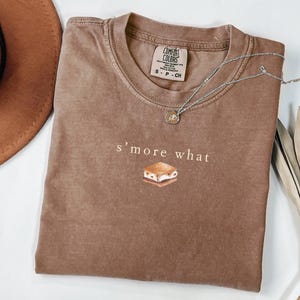 May include: A brown Comfort Colors t-shirt with the text "s'more what" and a graphic of a s'more. A silver necklace with a pendant is draped across the shirt. The shirt is folded and ready to wear.