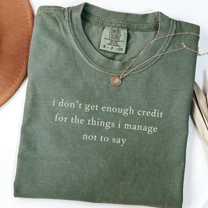 May include: Olive green t-shirt with the text "i don't get enough credit for the things i manage not to say" in white. A gold necklace with a pendant and a brown hat are also visible.