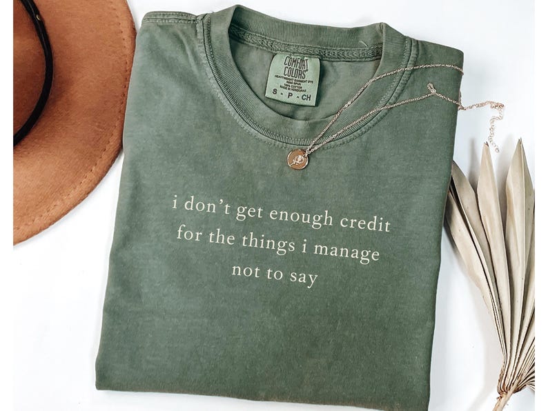 May include: Olive green t-shirt with the text "i don't get enough credit for the things i manage not to say" in white. A gold necklace with a pendant and a brown hat are also visible.