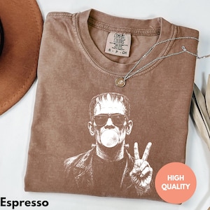 Puede incluir: Una camiseta marr&oacute;n de manga corta con un estampado gr&aacute;fico blanco de un personaje con gafas de sol, haciendo una burbuja y un signo de paz. La etiqueta interior dice Comfort Colors, Heavyweight Garment Dye, Ring Spun, 100% Cotton, Made in Honduras, S-P-CH.