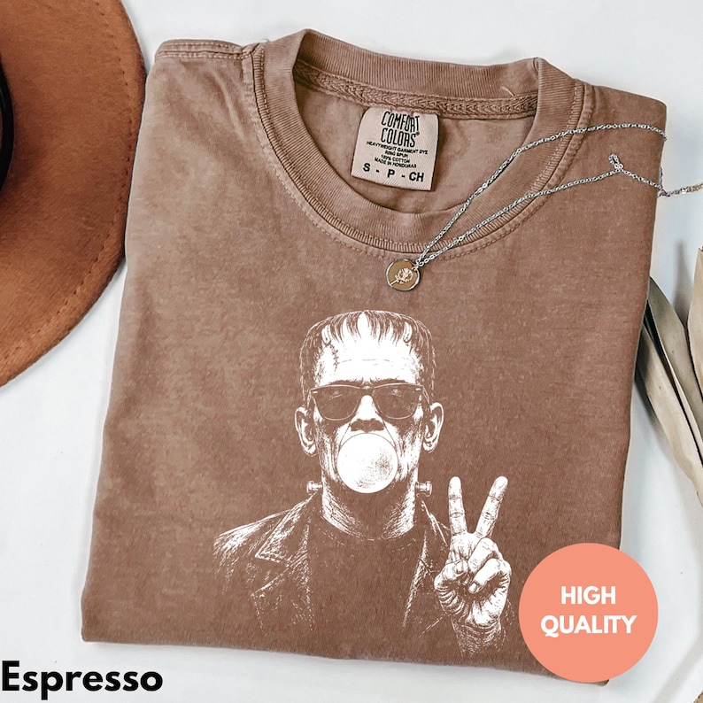 Puede incluir: Una camiseta marr&oacute;n de manga corta con un estampado gr&aacute;fico blanco de un personaje con gafas de sol, haciendo una burbuja y un signo de paz. La etiqueta interior dice Comfort Colors, Heavyweight Garment Dye, Ring Spun, 100% Cotton, Made in Honduras, S-P-CH.