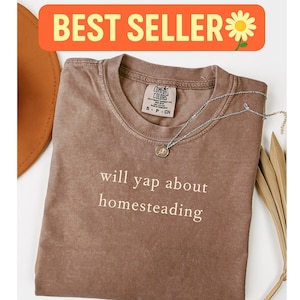 K&ouml;nnte beinhalten: Ein braunes Comfort Colors T-Shirt mit dem wei&szlig;en Text "will yap about homesteading". Eine silberne Halskette mit Anh&auml;nger liegt auf dem Shirt. Das Shirt ist gefaltet, mit einem Hut und getrockneten Bl&auml;ttern im Hintergrund. Die Worte "BEST SELLER" stehen in einem orangefarbenen Banner.