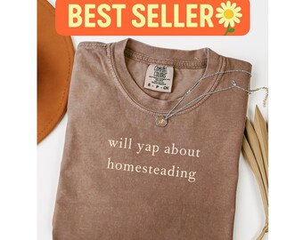 May include: A brown Comfort Colors t-shirt with the text "will yap about homesteading" in white. A silver necklace with a pendant rests on the shirt. The shirt is folded, with a hat and dried leaves in the background. The words "BEST SELLER" are in an orange banner.