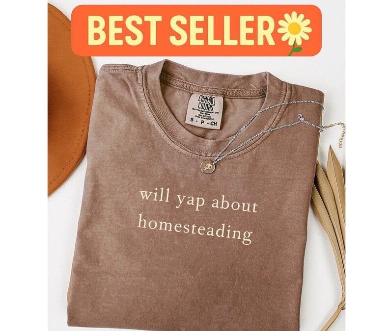 K&ouml;nnte beinhalten: Ein braunes Comfort Colors T-Shirt mit dem wei&szlig;en Text "will yap about homesteading". Eine silberne Halskette mit Anh&auml;nger liegt auf dem Shirt. Das Shirt ist gefaltet, mit einem Hut und getrockneten Bl&auml;ttern im Hintergrund. Die Worte "BEST SELLER" stehen in einem orangefarbenen Banner.