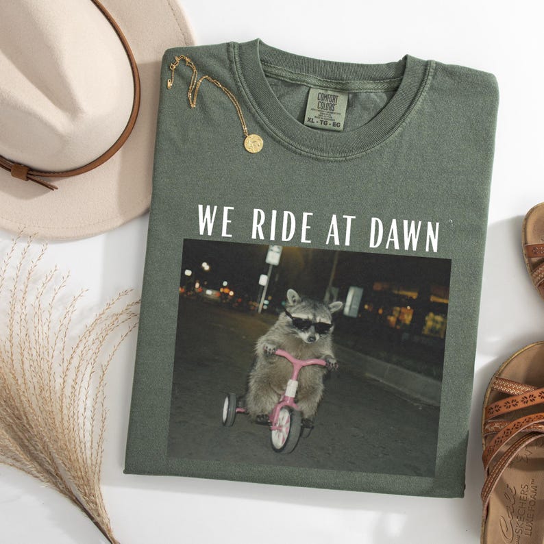 Puede incluir: Camiseta verde oliva con el texto "WE RIDE AT DAWN" sobre una foto de un mapache con gafas de sol montando en un triciclo rosa. Un collar dorado y un sombrero beige tambi&eacute;n est&aacute;n en la imagen.