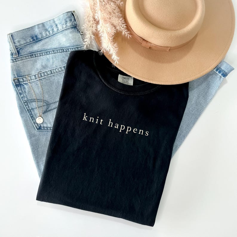 May include: A flat lay featuring a black t-shirt with the text "knit happens" in white, light blue jeans, a tan hat with a brown band, a gold necklace, and dried flowers. The t-shirt is the focal point of the image.