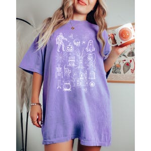 May include: A lilac t-shirt with a white Halloween graphic. The design includes a mummy, pumpkin, skull, ghost, spiderweb, Frankenstein, werewolf, skeleton, cat, haunted house, and cauldron. A white and orange mug with a sun design is held in the hand.