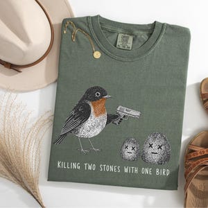 Puede incluir: Una camiseta verde oliva con el gr&aacute;fico de un p&aacute;jaro sosteniendo una pistola, apuntando a dos piedras con ojos en forma de X. El texto debajo del gr&aacute;fico dice: "KILLING TWO STONES WITH ONE BIRD." La camiseta tiene una etiqueta Comfort Colors.