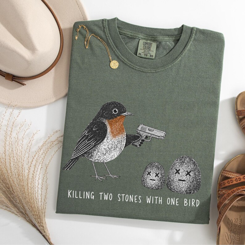Puede incluir: Una camiseta verde oliva con el gr&aacute;fico de un p&aacute;jaro sosteniendo una pistola, apuntando a dos piedras con ojos en forma de X. El texto debajo del gr&aacute;fico dice: "KILLING TWO STONES WITH ONE BIRD." La camiseta tiene una etiqueta Comfort Colors.