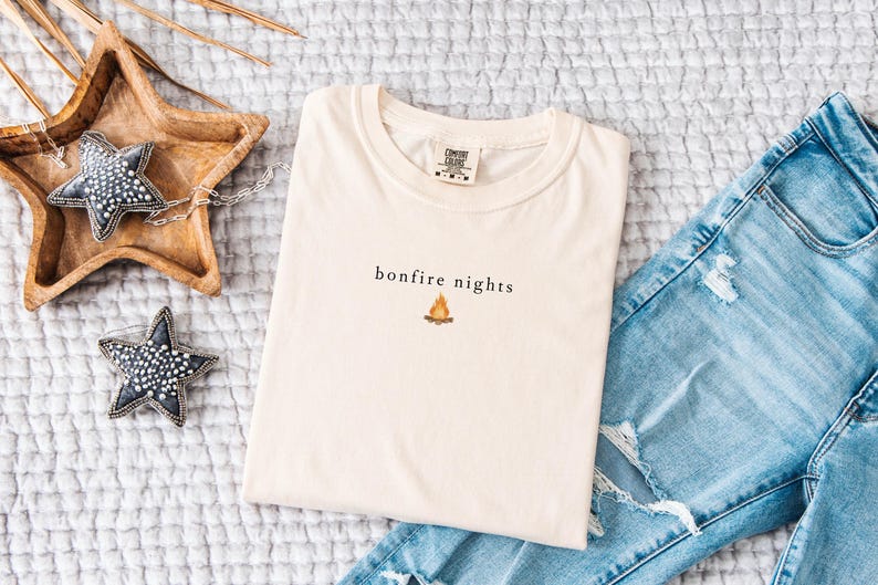 May include: Cream-colored t-shirt with the words "bonfire nights" and a small campfire graphic. The shirt is folded and placed on a textured surface next to a pair of blue jeans and decorative star-shaped objects.