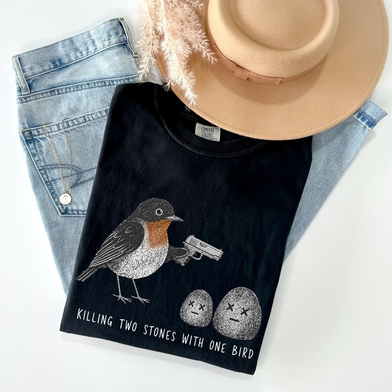 Puede incluir: Una camiseta negra con un gr&aacute;fico de un p&aacute;jaro sosteniendo una pistola, dos piedras con ojos en forma de X y el texto "KILLING TWO STONES WITH ONE BIRD". La camiseta se superpone a unos vaqueros azul claro, un sombrero beige y un collar dorado.