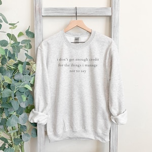 May include: A light gray sweatshirt with the text "I don't get enough credit for the things I manage not to say" in black. The sweatshirt is hanging on a wooden hanger against a white background.