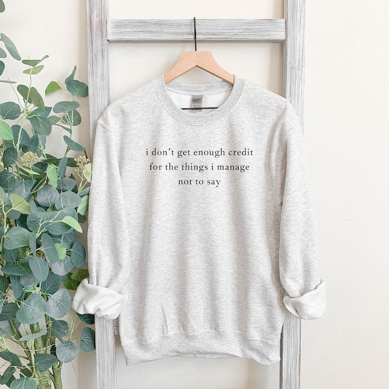 May include: A light gray sweatshirt with the text "I don't get enough credit for the things I manage not to say" in black. The sweatshirt is hanging on a wooden hanger against a white background.