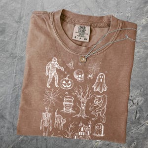 May include: A brown Comfort Colors t-shirt with a crew neck, featuring a white Halloween-themed graphic. The design includes a mummy, pumpkin, skull, spiderweb, ghost, Frankenstein, cat, haunted house, and a RIP tombstone. A silver necklace with a pendant is also present.