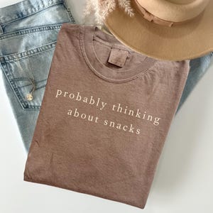 Pode incluir: Uma camiseta marrom com o texto "probably thinking about snacks" em letras brancas. A camiseta está dobrada em cima de um par de jeans azul claro, com um colar e um chapéu bege no fundo.