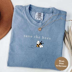 May include: A blue t-shirt with the phrase "save the bees" and a bee graphic. The shirt has a crew neck and short sleeves. A gold necklace with a pendant is on the shirt.