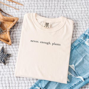 May include: Cream-colored t-shirt with the text "never. enough. plants" in black, paired with light blue distressed jeans. Decorative wooden star tray and beaded star ornaments complete the casual, plant-themed ensemble.