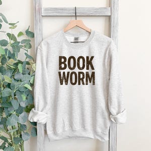 May include: A light gray sweatshirt with the words "BOOK WORM" printed in a dark brown, textured font. The sweatshirt is hanging on a wooden hanger against a white background.
