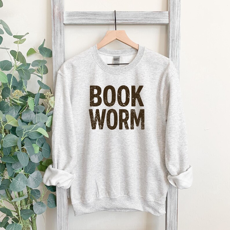 May include: A light gray sweatshirt with the words "BOOK WORM" printed in a dark brown, textured font. The sweatshirt is hanging on a wooden hanger against a white background.