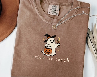 Puede incluir: Una camiseta marrón Comfort Colors con un fantasma de Halloween con un sombrero de bruja y sosteniendo una vela y una calabaza. El texto "trick or teach" está impreso debajo. Un collar plateado está sobre la camiseta.