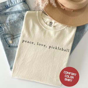 May include: Cream-colored t-shirt with the words "peace, love, pickleball" in black. The shirt is paired with blue jeans, a necklace, and a tan hat. A red circle with the words "Comfort Color Shirt" is in the bottom right corner.