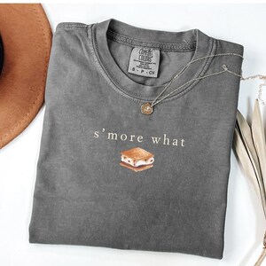 May include: A gray Comfort Colors t-shirt with the text "s'more what" and a graphic of a s'more. A gold pendant necklace and a brown hat are also visible. The shirt is a casual style.