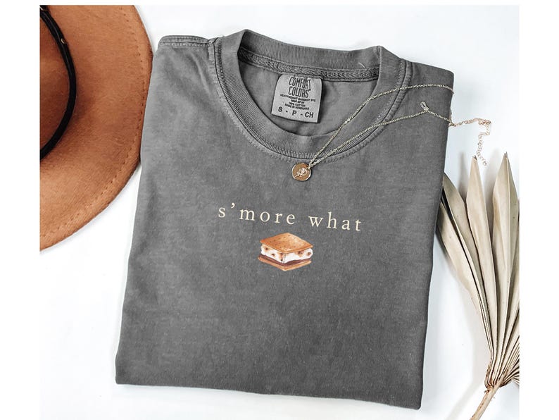 May include: A gray Comfort Colors t-shirt with the text "s'more what" and a graphic of a s'more. A gold pendant necklace and a brown hat are also visible. The shirt is a casual style.