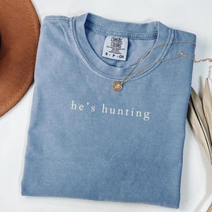 May include: A blue Comfort Colors t-shirt with the phrase "he's hunting" in white script. A gold necklace with a circular pendant rests on the shirt. A brown hat and dried leaves are in the background.