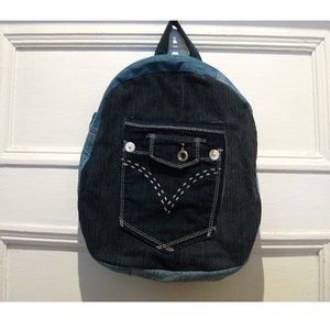 Peut inclure: Un sac à dos en jean bleu avec une grande poche sur le devant. La poche est ornée de surpiqûres blanches et d'un bouton argenté.