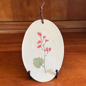 May include: Oval ceramic wall hanging with a floral design. The off-white plaque features a red flower and green leaf illustration. A brown leather cord is attached for hanging. The plaque is displayed on a black metal stand.