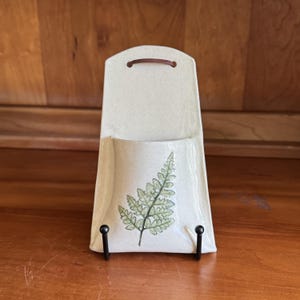 May include: A wall-mounted ceramic pocket planter with a speckled off-white glaze and a green fern leaf illustration on the front. The piece features a curved top edge with a brown leather hanging strap and is displayed on a black metal stand.
