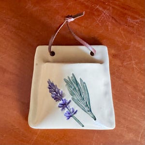 May include: Cream-colored ceramic wall pocket with a lavender and rosemary design. The pocket has a small opening at the top and a brown leather strap for hanging. The lavender is purple, and the rosemary is green.