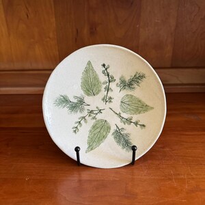 May include: A round, off-white ceramic plate featuring a green botanical design of leaves and sprigs. The plate is supported by a black metal stand, set against a warm wooden background. The design includes various leaf shapes.