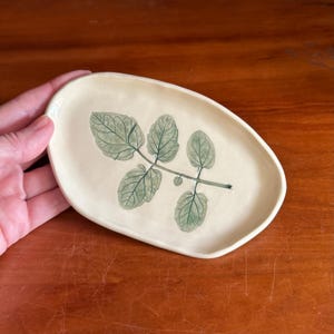 May include: Cream-colored ceramic oval dish with a hand-painted botanical design. The design features a green leafy branch with detailed veining. The dish has a slightly irregular shape, adding to its handmade charm. Ideal for serving small portions or as a decorative accent.