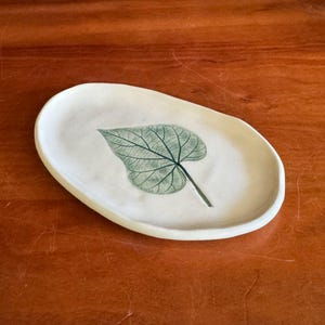 May include: Off-white ceramic oval dish with a green leaf design. The leaf has detailed veining and a dark green stem. The dish is set against a warm brown wooden surface.