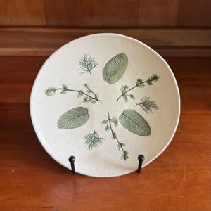 May include: A round, off-white ceramic plate featuring a botanical design. The plate is decorated with green leaf and sprig illustrations, including sage and dill. The plate is displayed on a black metal stand, set against a wooden background.