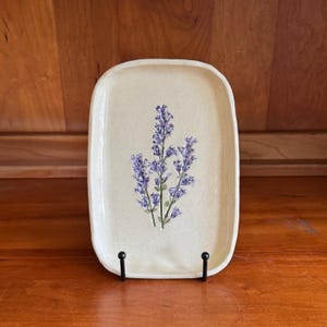 May include: A rectangular ceramic tray with rounded corners, featuring a speckled off-white glaze and a central botanical illustration of purple lavender sprigs with green stems. The tray is held upright by a black metal display stand.