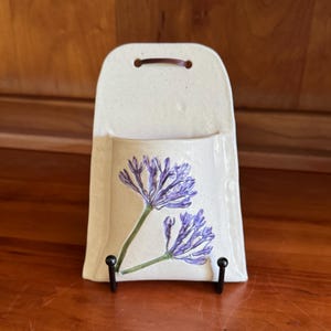 May include: A cream-colored ceramic wall pocket with a speckled finish and a brown leather hanging strap. The front features a hand-painted illustration of two purple agapanthus flowers with green stems, positioned vertically on the flat surface.