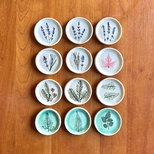 May include: A collection of twelve small, round ceramic dishes. Each dish features a hand-painted botanical design, including lavender, leaves, and a maple leaf. The dishes are arranged on a wooden surface, showcasing the variety of designs and colors.