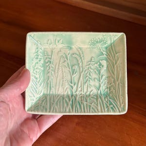 May include: A rectangular ceramic dish with a raised botanical design. The dish features a light green background with white embossed plants and flowers. The edges are slightly raised, and the dish is held by a hand.