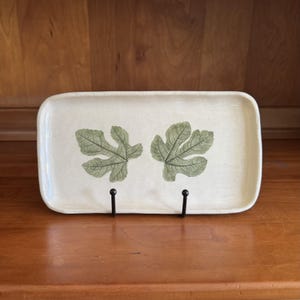 May include: A rectangular ceramic tray with a speckled off-white glaze featuring two green fig leaf imprints centered on the surface. The tray has slightly raised edges and is supported by a black metal stand, showcasing a smooth finish.