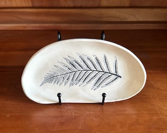 May include: Cream-colored ceramic oval dish with a black fern leaf design. The dish is supported by a black metal stand. The fern leaf is detailed with fine lines and dots, creating a naturalistic aesthetic.