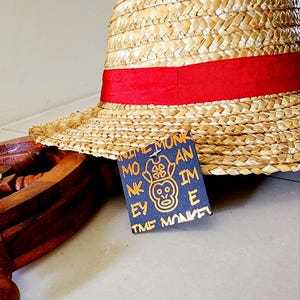 May include: A straw hat with a red band and a black tag that says "Anime Monkey" with a monkey graphic in gold. The tag also says "Monkey D. Luffy" in gold.