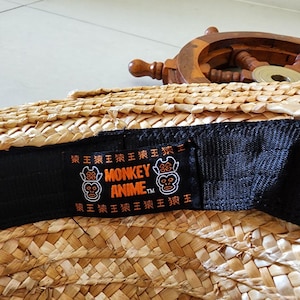 May include: A black hat with a woven straw crown and a black adjustable strap. The strap has a label with the text "MONKEY ANIME TM" and two orange monkey faces.