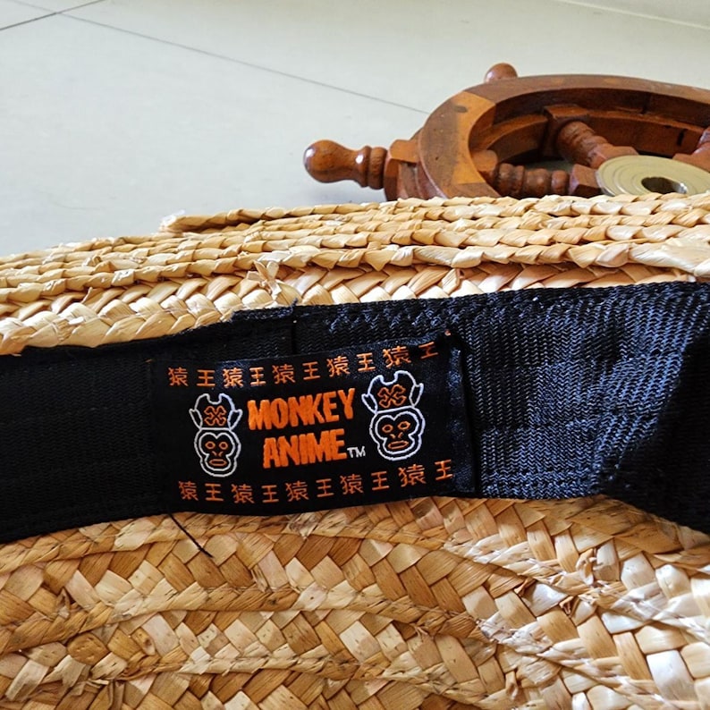 May include: A black hat with a woven straw crown and a black adjustable strap. The strap has a label with the text "MONKEY ANIME TM" and two orange monkey faces.