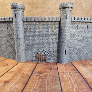 Könnte beinhalten: Eine graue Miniatur-Burgmauer mit zwei zylindrischen Türmen und einem zentralen Bogeneingang. Die Mauer besteht aus rechteckigen Blöcken mit Zinnen an der Oberseite. Die Türme haben kleine Fenster. Das Schloss ist ein Modell.