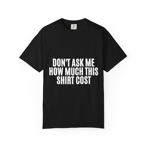 以下が含まれることがあります： 白い文字で「DON'T ASK ME HOW MUCH THIS SHIRT COST」と書かれた黒いTシャツ。Tシャツは半袖で、クルーネックです。テキストはシャツの前面の中央に配置されています。
