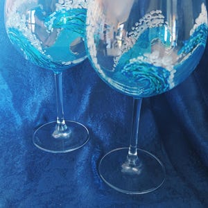 Op de afbeelding: Twee heldere wijnglazen met lange stelen. Elk glas is met de hand beschilderd met een blauw en wit golfpatroon. De glazen staan tegen een blauwe achtergrond.