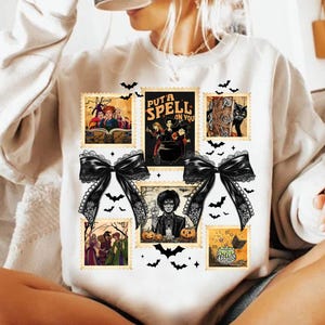 Peut inclure: Sweat-shirt blanc orné d'un collage de timbres-poste sur le thème d'Halloween. Les timbres représentent des sorcières, un chat noir et la phrase "PUT A SPELL ON YOU". Deux grands nœuds noirs et des silhouettes de chauves-souris complètent le design.