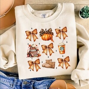 Könnte beinhalten: Ein cremefarbener Pullover mit einem Herbst-Motiv. Das Design zeigt einen Stapel Bücher, einen Kürbis, eine Tasse Kaffee und vier braune Schleifen.